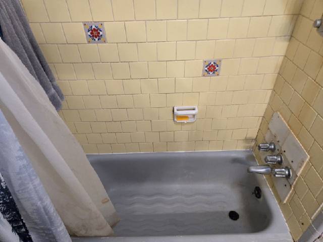 Bathtub surround with cream square wall tile and a temporary backing board around the faucet during repair