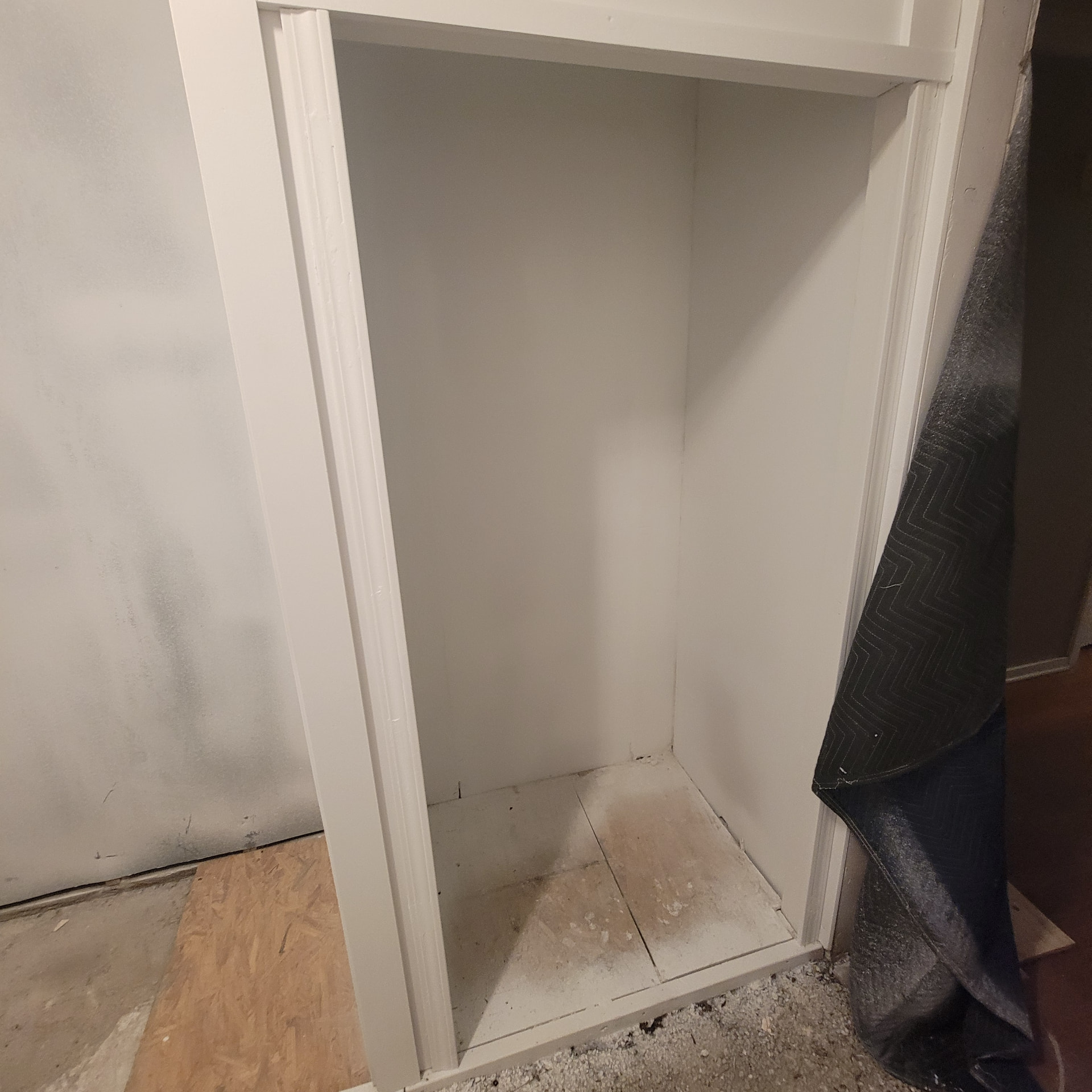 Newly framed and painted built-in bathroom storage niche with off-white trim, unfinished floor base and a protective moving blanket to the side