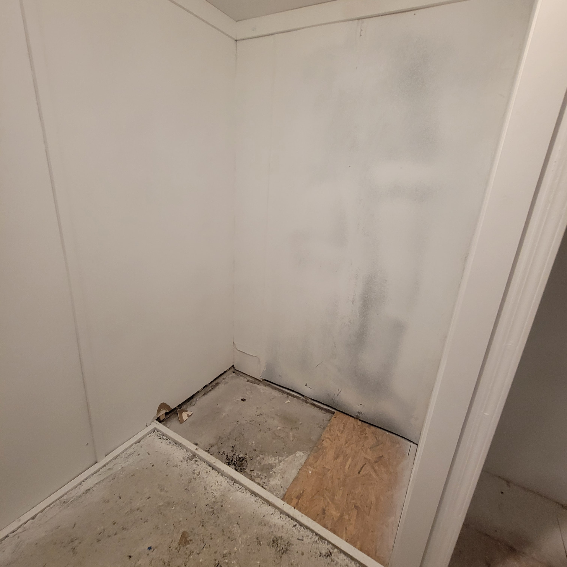 Bathroom floor stripped for new flooring with exposed gray concrete subfloor and an OSB patch, white walls and loose trim on the floor