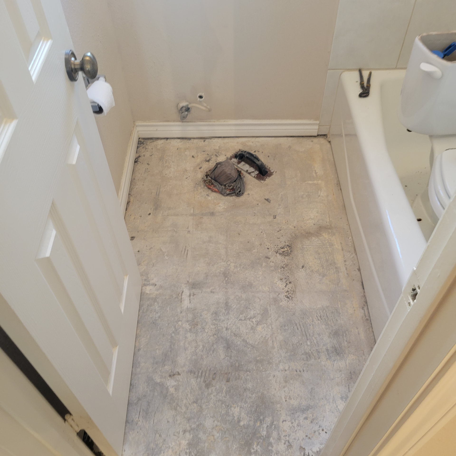 Bathroom mid-demolition with flooring removed to the concrete subfloor, white tub still in place, toilet tank set inside the tub and a plugged toilet flange